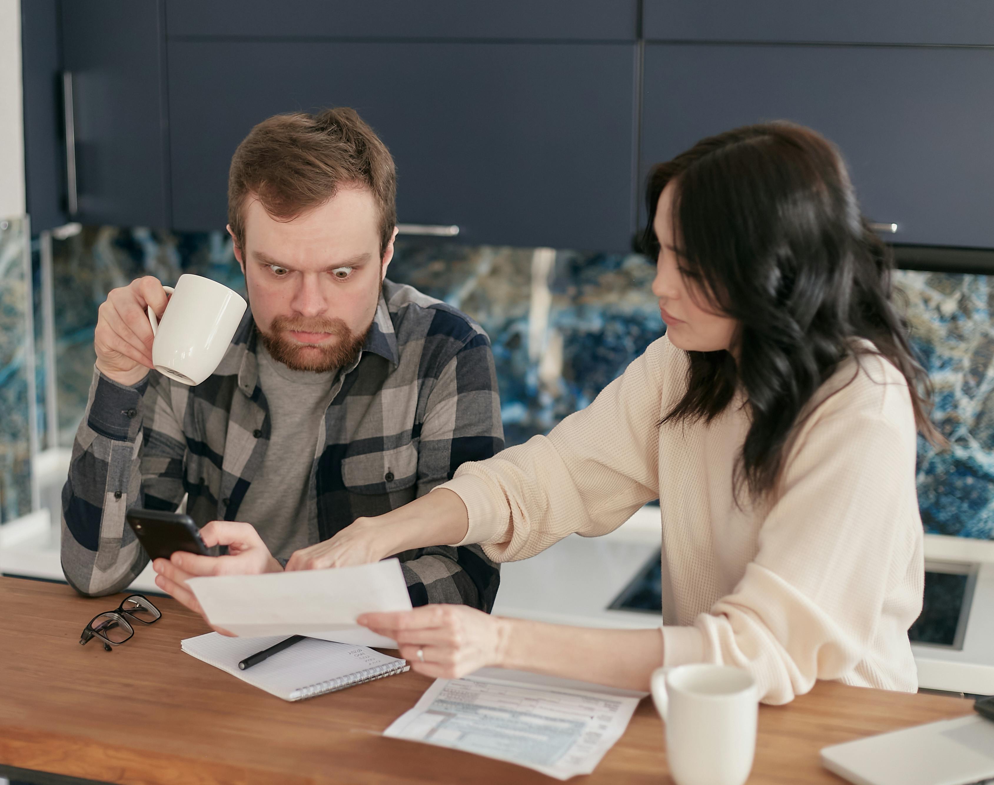 A couple reviewing bills at a kitchen table while one of them looks at a phone, illustrating monthly-payment stress.